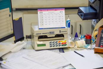 Foto de uma impressora de cheques. No centro da imagem está uma máquina impressora de cheques branca com botões coloridos. Em cima dela, há um calendário encadernado em espiral que mostra o mês de outubro de 2023. Do lado esquerdo, há um monitor de computador e do lado direito há objetos de papelaria. Todos objetos estão sobre uma mesa de escritório com papéis de rascunho e documentos.