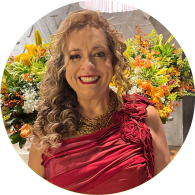 Foto de uma mulher branca, com cabelos loiros, longos e ondulados, sorrindo. Ela usa batom vermelho, colares dourados e vestido vermelho com rosas de tecido sobre um dos ombros e sem alça do outro lado lado. Atrás dela há arranjos de flores amarelas e laranjas, uma parede cinza e uma luminária.