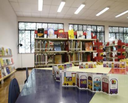 Foto de cartões sanfonados com diversas ilustrações coloridas sobre a mesa de uma biblioteca. Ao fundo, há prateleiras cheias de livros e grandes janelas.