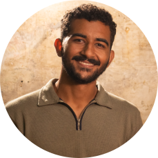 Foto de um homem sorrindo. Ele é negro, com cabelo curto cacheado castanhos escuros, bigode e barba castanhos e olhos pretos. Veste camisa polo cinza, com detalhes em preto. Ao fundo está uma parede de cimento manchada. A iluminação é amarelada.