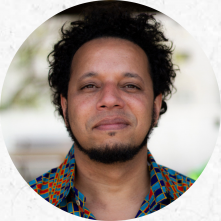 Foto redonda de um homem sorrindo de leve. Ele é negro, tem cabelos curtos e escuros, olhos escuros e barba. Ele veste uma camisa estampada com quadrados azuis claro e escuro e laranja.