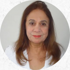 Foto do rosto de uma mulher de pele clara, cabelos castanhos longos abaixo do pescoço e idosa. Ela olha para a câmera, sorri levemente e usa uma blusa branca.