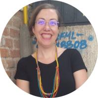 Foto de uma mulher branca sorrindo. Ela tem cabelos lisos e castanhos, que estão presos, veste blusa preta de mangas curtas, usa colar com miçangas amarelas, vermelhas e azuis e óculos de armação redonda e transparente. Ao fundo, à esquerda da foto,  há uma parede de tijolos e, à direita, uma parede  com escritos em tinta azul.