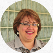 Foto de mulher branca sorrindo. Ela tem cabelos castanhos claros, curtos e lisos, usa óculos de armação fina, brincos brancos e camisa social cinza.