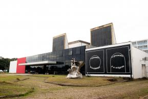 Foto da fachada do Espaço das Artes. Ela é dividida em quatro partes. Da esquerda para a direita, a primeira é uma parede branca e a segunda é vermelha. A terceira, que é a mais comprida, tem vidros pretos reflexivos na parede e uma cobertura branca com bancos embaixo. A última parte é uma parede com a pintura de um retângulo preto e as frases “o passado é o começo” e “o presente é a origem” em branco. Na frente do prédio, há uma escultura de metal e um gramado no chão.