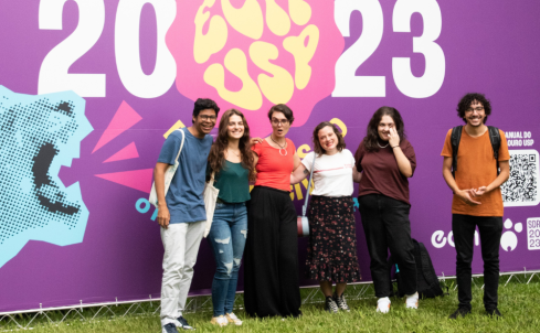 Seis pessoas, dois homens e quatro mulheres, estão de pé, abraçados um do lado do outro, exceto por um deles, mais à direita, enquanto posam para uma foto. Ao fundo, há um banner de divulgação da Semana de Recepção aos Calouros, nas cores roxa, rosa, amarelo, azul e branco. Nele está escrito, ECA USP, em amarelo, sobre um fundo Rosa, e 2023, em branco. O desenho de um leão rugindo, em azul, está no canto esquerdo