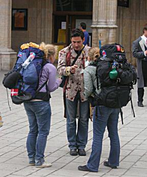 Foto de duas jovens conversando com um homem. Ele está no centro da imagem, veste uma camisa com estampa florida e calça jeans. Ele segura uma caneta e parece estar orientando as moças. A jovem da esquerda tem cabelos cacheados e loiros, ela usa um casaco roxo, calça jeans, e mochila azul. A jovem da direita tem cabelo castanho com mechas loiras, usa uma camisa xadrez, calça jeans e mochila preta.