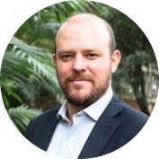 Foto de um homem branco com um ligeiro sorriso. Ele é careca, tem barba e bigode castanhos, olhos castanhos, veste camisa branca e terno azul-marinho. Atrás dele, folhas de árvores levemente desfocadas. 