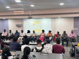 Foto em uma sala de aula. Em primeiro plano, de costas para a câmera, estão diversos jovens sentados. Na frente da turma, está uma fileira com 11 pessoas sentadas. Atrás delas, há uma projeção de um cartaz de divulgação do concurso. 