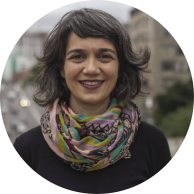 Foto de mulher branca sorrindo. Ela tem  olhos castanhos, cabelos grisalhos e ondulados de tamanho médio, veste uma blusa preta e usa um lenço colorido no pescoço. Atrás dela há um cenário urbano com ruas, carros e prédios desfocados.