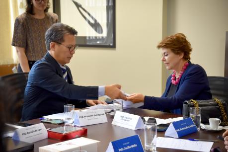  Foto de duas pessoas sentadas em uma mesa de reuniões. À esquerda, um homem amarelo, vestindo paletó azul marinho e gravata, e uma mulher branca, de cabelos curtos, lisos e castanhos, vestindo um blazer azul marinho e colar rosa. Com uma das mãos, ele entrega a ela um documento. Sobre a mesa, estão papéis, copos de água e alguns objetos. Uma mulher em pé os observa. Ao fundo, uma parede bege.