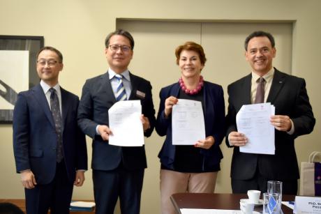  Foto de quatro pessoas, de pé, posando e sorrindo. Da esquerda para direita estão: dois homens asiáticos, vestindo terno e gravata, uma mulher branca, de cabelos curtos, lisos e castanhos, vestindo um blazer azul marinho e um homem pardo, vestindo terno e gravata. Dois homens e a mulher seguram com as duas mãos uma folha de papel cada com as assinaturas do convênio. Ao fundo, uma parede bege. 