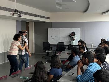 Foto de pessoas na sala de aula. À esquerda da imagem, uma mulher está em pé conduzindo um homem, que está com os olhos vendados. À direita estão diversos jovens sentados e apoiados em mesas observando a cena. Ao fundo, está um quadro branco e uma pessoa tirando fotos com uma câmera profissional.