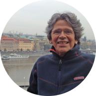 Foto de homem sorrindo de frente para a câmera. Ele é branco e tem cabelos lisos, curtos e grisalhos, usa óculos de armação transparente e veste casaco azul com detalhes vermelhos. Ao fundo, um rio e edifícios. 