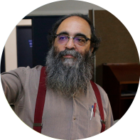 Foto de um homem de pele clara, calvo, de cabelos grisalhos, barba longa e grisalha. Ele veste uma camisa cinza com suspensórios vermelhos. O fundo mostra tratar-se de  um ambiente fechado.