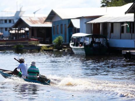 Foto de dois homens em um rio, dentro de um pequeno barco motorizado. Atrás deles estende-se um rastro na água, deixado pelo movimento das hélices do motor. Ao lado deles encontram-se algumas habitações de palafita.