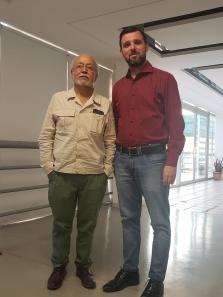 Foto de Wellington ao lado do professor Gómez. À esquerda, está o professor, de pé e com as mãos nos bolsos da calça. É um homem branco, de barba e cabelos grisalhos e veste uma camisa bege, calça verde e sapatos marrons. Ao seu lado está Wellington, de pé com as mãos ao lado do corpo. É um homem branco, de cabelos castanhos e barba castanha com algumas partes grisalhas, veste camisa social vermelha, calça jeans e sapatos pretos.