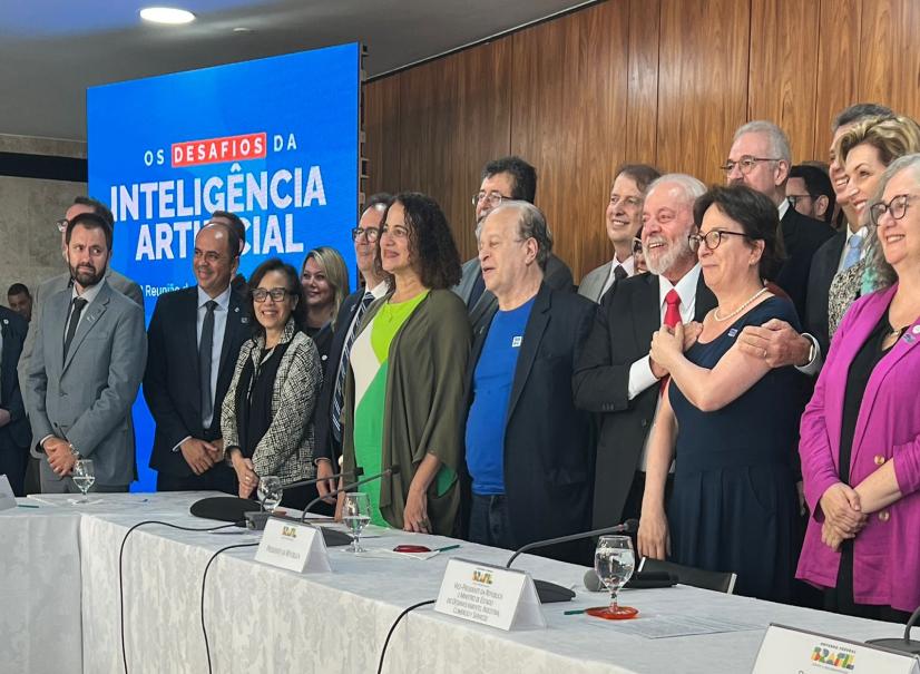 Foto de pessoas diversas em pé, sorrindo e posando para o lado esquerdo da imagem. Elas aparentam ter meia idade e são majoritariamente brancas, vestem roupas formais e estão diante de uma bancada, sobre a qual estão microfones, taças com água e placas de identificação. Do lado esquerdo da imagem, atrás de algumas pessoas, há um banner azul, no qual está escrito “os desafios da Inteligência Artificial”. Ao fundo, uma parede de madeira. A professora Roseli Fígaro, uma mulher branca de cabelos lisos, curtos e escuros, se encontra do lado direito da imagem abraçada com o Presidente Lula, um homem idoso de pele branca, barba e cabelos curtos brancos.