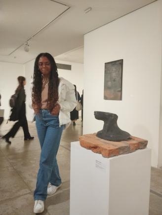 Na foto, uma mulher negra com as mãos nos bolsos olha para uma escultura em uma sala de exposição. Ela tem tranças longas e castanhas, usa óculos, veste calça jeans, camiseta rosa, casaco e calça tênis brancos. A escultura, à frente da imagem, é a parte inferior de um rosto e está sobre um expositor retangular branco. Ao fundo, um quadro pendurado na parede e pessoas caminhando.