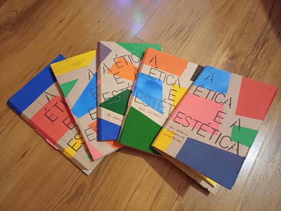 Foto de cinco livros cartoneros parcialmente sobrepostos formando um semicírculo. Todos possuem capas de papelão com formas geométricas coloridas e o título “A ética e a estética” em fonte preta. Estão sobre um piso de madeira.