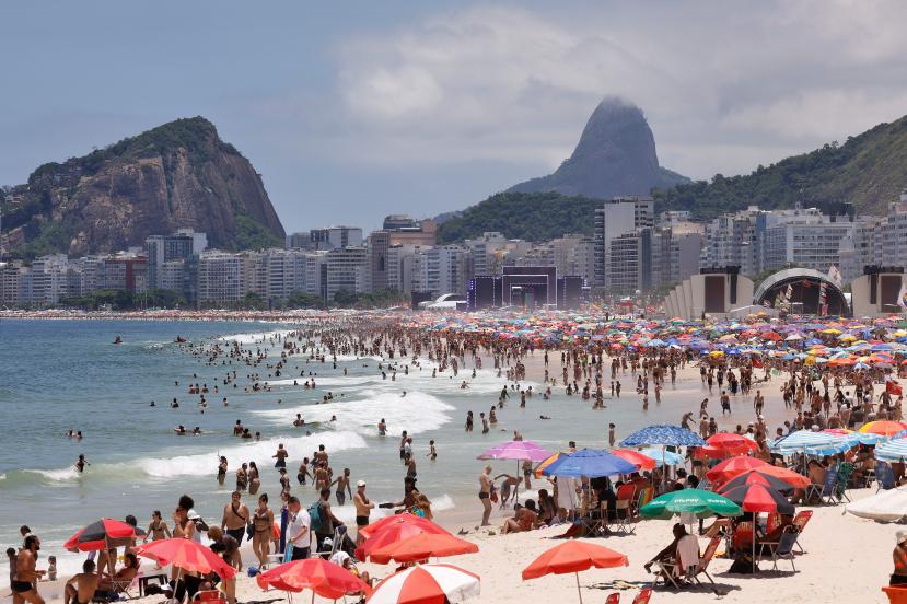 Foto de uma praia com centenas de pessoas. O mar é azulado e a areia está repleta de guarda-sóis coloridos e pessoas sentadas em cadeiras de praia, em pé ou na água. Ao fundo da faixa de areia, há duas estruturas de palcos musicais. Atrás delas, há dezenas de prédios modernos que cobrem parcialmente montanhas. O céu está azul com poucas nuvens.
