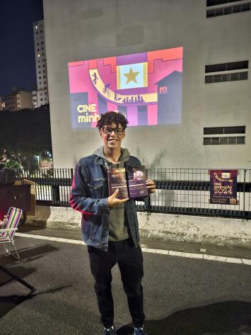 Foto de rapaz jovem negro de cabelos escuros, cacheados e curtos. Ele usa óculos e segura um folder. Ele está de jaqueta jeans sobre blusa verde e de calças escuras. No fundo da imagem, uma projeção sobre a parede de um prédio com o escrito “Cine Minhocão”.