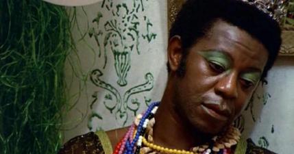 Cena do filme. Pessoa negra, cabelo crespo baixo, aparentemente uma coroa prateada na cabeça. A pessoa usa sombra verde, batom vermelho e vários colares. Está encostada na parede e olha para o lado aparentemente reflexiva. Na parede há desenhos feitos com linhas verdes