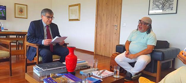 Fotografia de dois homens sentados em uma sala fechada. Em primeiro plano, há uma mesa de centro com alguns livros. À esquerda, está sentado um homem branco de óculos e cabelo grisalho. Ele veste terno azul e gravata vermelha e  olha para algumas folhas de papel em sua mão. À direita, um homem negro, de óculos e tranças curtas. Ele veste uma boina bege, camiseta azul e calça branca enquanto fala e olha para o homem ao seu lado. Em segundo plano, há uma porta de madeira, alguns quadros na parede e parte de uma mesa.