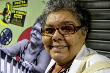 Foto na altura do ombro de uma mulher de pele negra com cabelo curto e grisalho e óculos de aro retangular. Ela sorri olhando para a câmera. Ao fundo, um cartaz com fundo verde e vermelho com algumas ilustrações e uma imagem da mesma mulher em preto e branco.