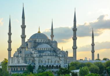 Foto colorida em ambiente externo, de uma grande mesquita com cinco torres pontiagudas ao seu redor.