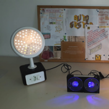 Foto de três luminárias acesas feitas com materiais reciclados. Da esquerda para a direita, uma que parece a ducha de um chuveiro em cima de uma caixa preto com um espelho de interruptor e tomada, com pontos de luz amarela saindo pelos buracos.