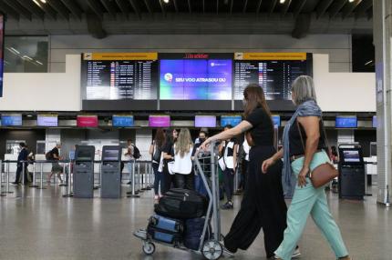 Foto de um aeroporto. Duas mulheres caminham enquanto olham para o painel de voos posicionado acima dos guichês de atendimento. Uma das mulheres é branca, tem cabelos longos escuros, veste camiseta e calça pretas e empurra um carrinho com malas pequenas. A outra mulher é parda, tem cabelos grisalhos na altura dos ombros, veste camiseta preta, calça azul claro e carrega uma bolsa pequena marrom. Não é possível ver o rosto delas.