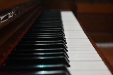 Foto das teclas de um piano, partindo do primeiro plano da imagem para um fundo mais desfocado. As teclas são brancas e pretas e a madeira do piano é avermelhada.
