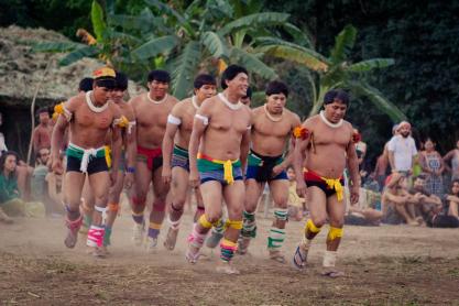 Foto de vários homens indígenas performando uma dança típica. Todos vestem uma espécie de colar branco, algo que parece um shorts curto com faixas coloridas amarradas na cintura, bem como outras faixas de diversas cores amarradas na altura do tornozelo e um pouco abaixo do joelho. Eles dançam sobre um chão de terra batida. Ao fundo, é possível distinguir uma habitação, palmeiras e outras pessoas, que parecem assistir a dança. 