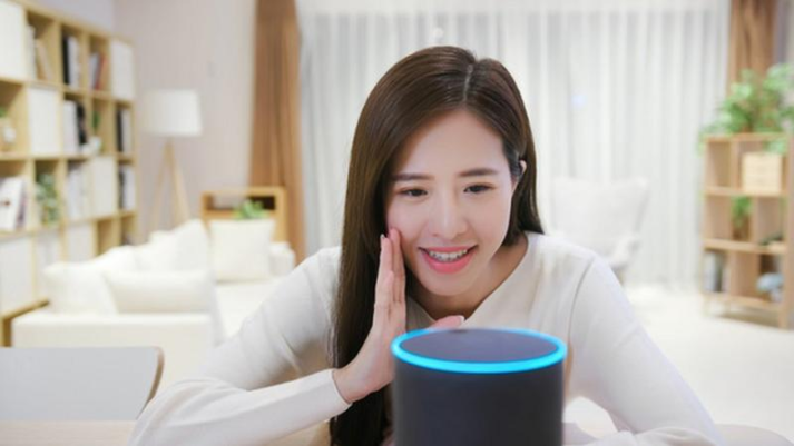 Foto de uma mulher amarela de cabelos escuros e compridos que coloca a mão próxima à boca interagindo com a assistente virtual Alexa da empresa Amazon, representada por um cilindro preto com a borda superior iluminada na cor azul. Ao fundo, há uma sala de estar com sofá, luminária, estantes e vasos com plantas. Predomina a cor branca.