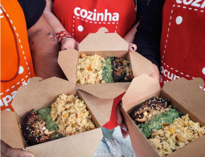 Foto de três caixas de papelão com comida. Cada uma delas é carregada por uma pessoa diferente, e as três usam um avental vermelho com o logo do projeto. Dentre outros alimentos, é possível identificar arroz e salada na refeição.