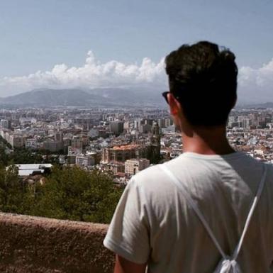 homem branco, com uma camisa clara e uma mochila, está de costas, olhando para a paisagem urbana e céu com nuvens ao fundo