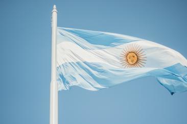 Foto colorida em ambiente externo, com a bandeira da Argentina em frente a um céu azul