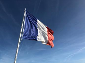 Foto colorida em ambiente externo, da bandeira da França ao vento com o céu azul de fundo