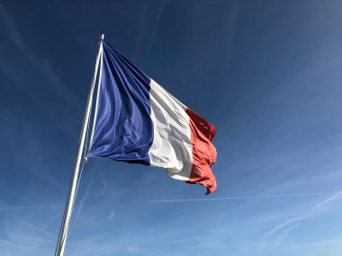 Foto colorida em ambiente externo, da bandeira da França balançando diante do céu cerúleo