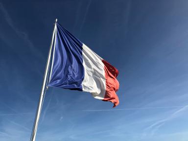 Foto colorida em ambiente externo, da bandeira da França balançando diante do céu azul