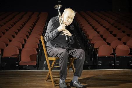 A foto destaca um homem branco, idoso, de bigode, careca, com cabelos brancos dos lados da cabeça. Ele está com roupas pretas, sentado em uma cadeira em cima de um palco, segurando um trompete apoiado no ombro. Ao fundo estão as cadeiras do teatro, que são vermelhas