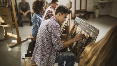 Foto colorida em ambiento interno, de um jovem rapaz desenhando em uma tela, com outros estudantes ao fundo