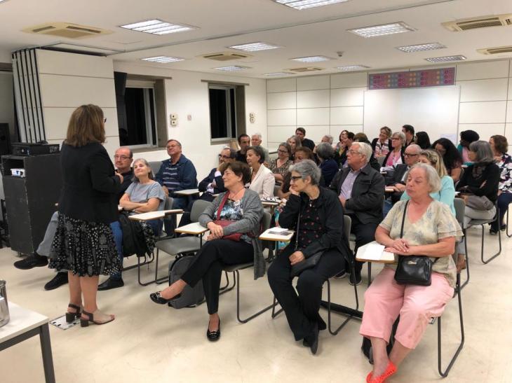 Fotografia colorida mostra aula do Programa USP 60+. Do primeiro plano até o plano de fundo, quase todas as carteiras estão ocupadas por homens e mulheres idosos. Alguns sorriem enquanto acompanham a explicação de uma professora, de costas para a câmera. 