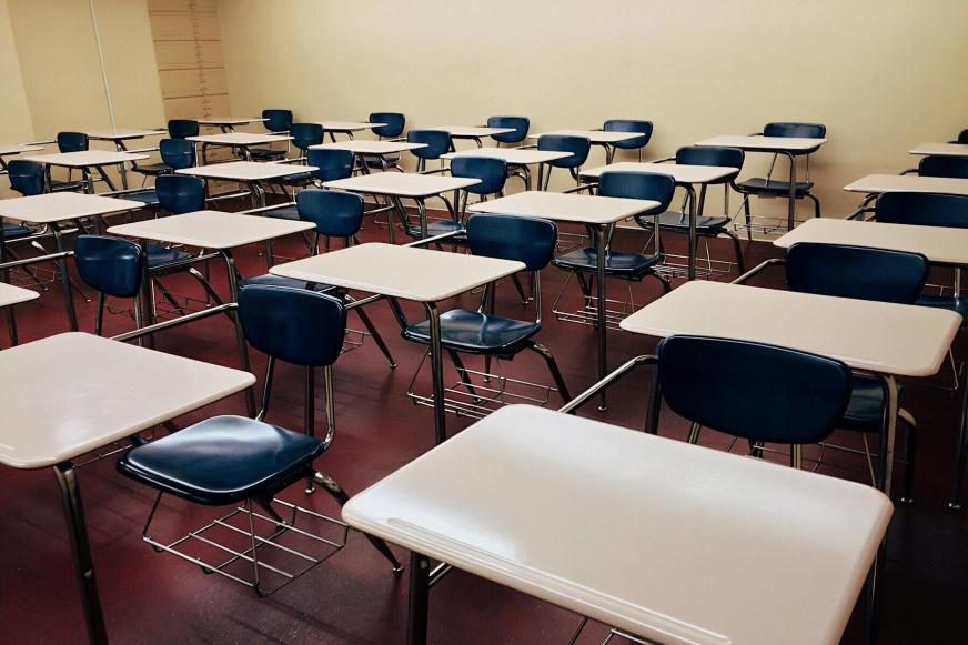  Sala de aula vazia de paredes amarelas claras e chão vermelho escuro. As mesas são brancas e as cadeiras azuis com a estrutura de suporte preta. Elas estão enfileiradas em 6 fileiras com 5 carteiras em cada.