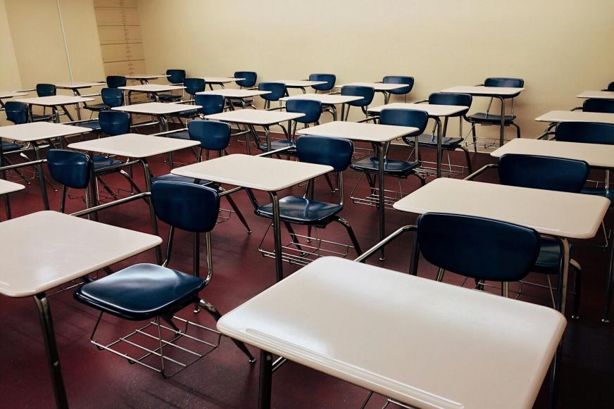 Sala de aula vazia de paredes amarelas claras e chão vermelho escuro. As mesas são brancas e as cadeiras azuis com a estrutura de suporte preta. Elas estão enfileiradas em 6 fileiras com 5 carteiras em cada.