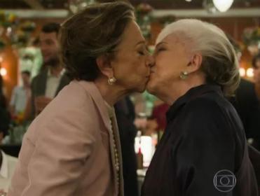 Foto de duas mulheres idosas se beijando nos lábios. Teresa está com uma roupa rosa, usando brincos e um colar e tem cabelo curto e castanho com mechas grisalhas. Estela está de roupa preta, brincos e tem cabelos curtos majoritariamente brancos.