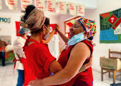 Foto de duas mulheres dançando. As duas são negras, estão com cabelos presos em faixas de tecido e usam o avental vermelho do projeto. Uma delas usa máscara e a outra a deixa pendurada na orelha, mostrando seu sorriso. Alguns cartazes aparecem desfocados presos nas paredes ao fundo.