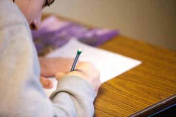 Foto colorida em ambiente interno, de uma pessoa debruçada sobre uma mesa escolar para fazer uma prova.