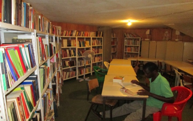 Foto da biblioteca da Universidade. Nas paredes, estão encostadas prateleiras de metal repletas de livros. Mesas de madeira formam um semicírculo no meio da sala. A maioria das cadeiras é de plástico. Um rapaz está sentado em uma delas. Ele é negro, tem cabelos curtos e escuros e veste uma camiseta verde. Ele está lendo um livro. O ambiente é escuro, apenas uma lâmpada amarela o ilumina.